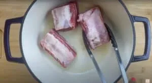 Beef short ribs being browned in a dutch oven with tongs, preparing for a delicious stew.